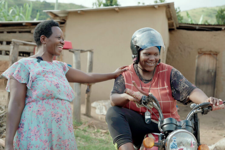 Uganda: A woman in a dress stands next to a woman motorcycle driver, her hand on her shoulder. They laugh togehter.